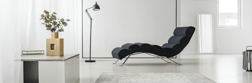 Black chaise lounge placed in white interior with metal lamp plant in gold vase and grey carpet